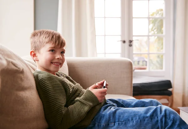 自宅でソファの上の若い男の子は携帯電話でゲームを楽しんでいます — ストック写真