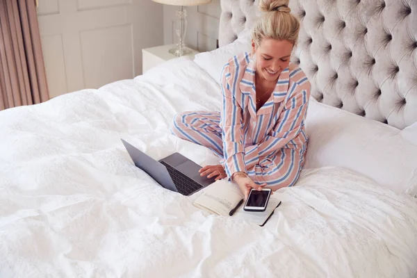 Businesswoman In Pyjamas Sitting On Bed With Laptop And Mobile Phone Working From Home In Lockdown