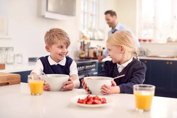 父が仕事の準備をするにつれてキッチンで学校の制服を着て2人の子供が朝食を食べる — ストック写真