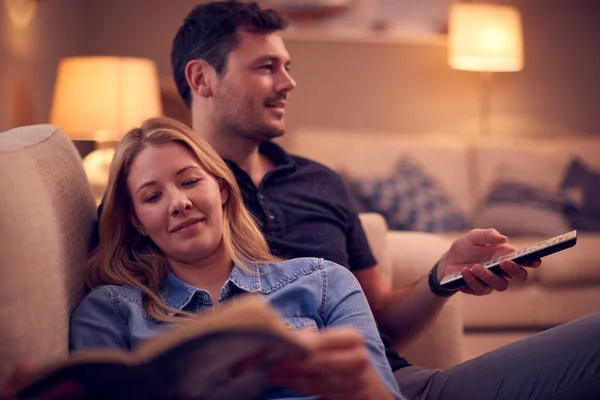 女性は本や男性の腕時計テレビを読むように自宅でソファでリラックスしたカップルの夜のショット — ストック写真