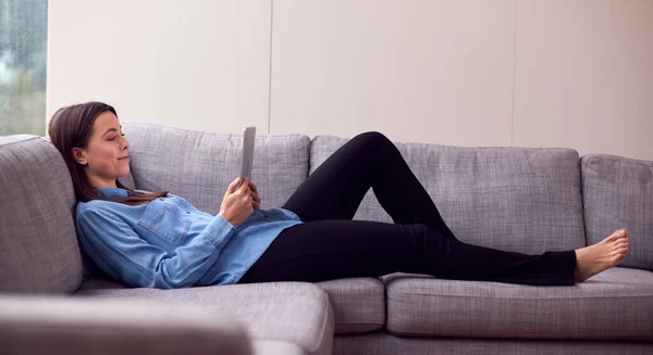 自宅で笑顔若い女性は デジタルタブレットを見てソファに横たわっています — ストック写真