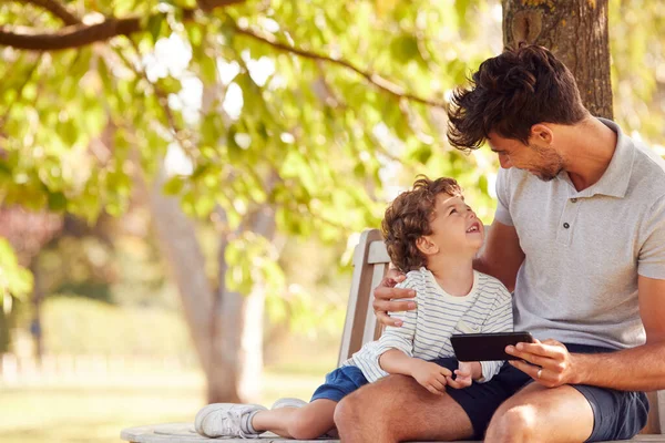 父亲坐在树下的公园长椅上 儿子一起看手机 — 图库照片