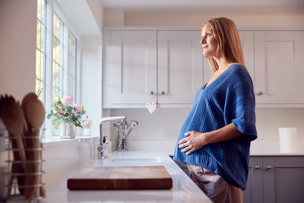 Uśmiechnięta Kobieta Ciąży Stojąca Przy Oknie Kuchennym Dotykająca Żołądka — Zdjęcie stockowe