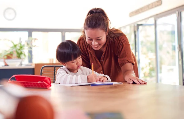 アジアの母は本で書く台所のテーブルで働く家庭の学校の息子を助ける — ストック写真