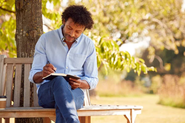 大人の男はノートに書く木の下の公園ベンチに座って — ストック写真