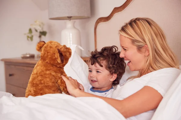 お母さんと若い息子が一緒にベッドルームでパイジャマを身に着けているかわいいテディベアのおもちゃと遊ぶ — ストック写真