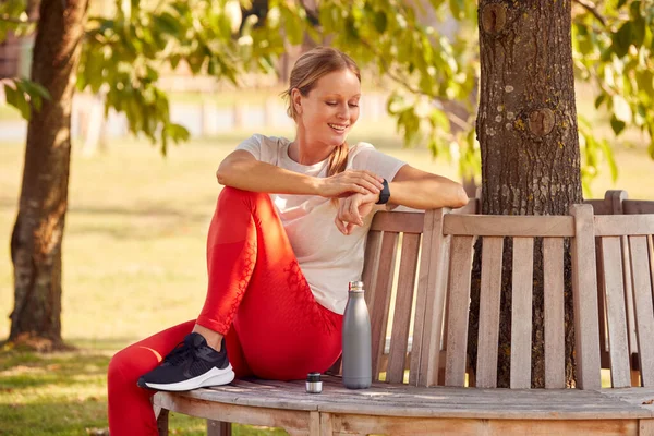 Frau Fitnessbekleidung Sitzt Auf Sitz Unter Baum Und Überprüft Aktivitätsmonitor — Stockfoto