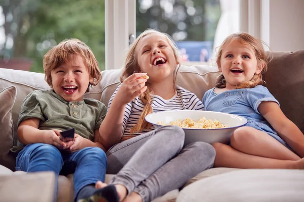 三个孩子坐在沙发上在家里笑着看电视吃爆米花 — 图库照片
