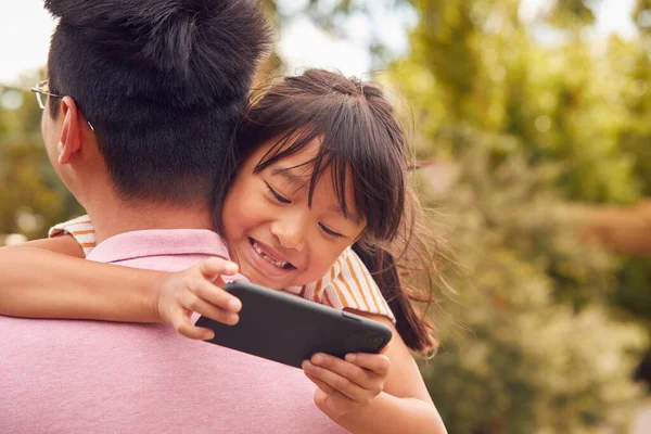 女の子は携帯電話で彼の肩の上に見えるように庭でアジアの父の冷笑の娘 — ストック写真