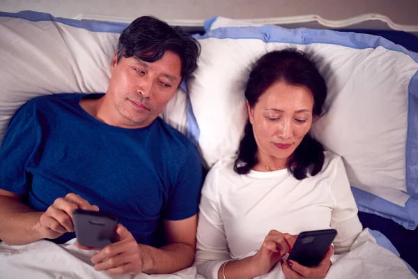 Зрелая Азиатская Пара Пижаме Лежащая Постели Смотрящая Мобильные Телефоны Вместе — стоковое фото