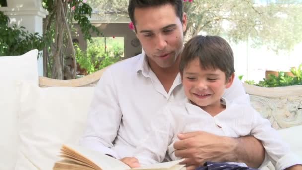 Отец и сын сидят в саду, читая книгу — стоковое видео