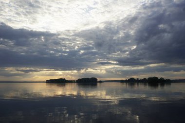      Sıcak bir yaz gününde gölün üzerinde gün batımı. Haziran gökyüzü ve güzel bulutlar. Ufuk ve yansıma. Suyun pürüzsüz yüzeyi.                           