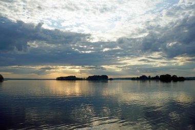 Sıcak bir yaz gününde gölün üzerinde gün batımı. Haziran gökyüzü ve güzel bulutlar. Ufuk ve yansıma. Suyun pürüzsüz yüzeyi.                               