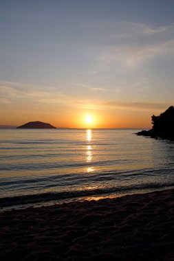 Ege Denizi üzerinde parlak kırmızı gün batımı. Güneş ve dağlar. Ufuk. Dalgalar ve parıltılar. Kumsal. Dikey çekim                               