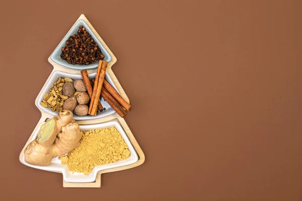 Cloves, cinnamon, nutmeg, cardamom and ginger for making ginger cookies in a box in the shape of a Christmas tree on a brown background with copy space. Concept of festive Christmas baking. Top view, flat lay.