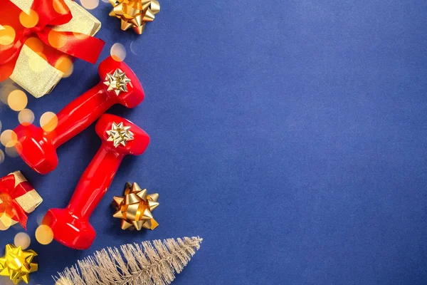 Cvičení, fitness a rozvoj konceptu šťastných Vánoc a šťastný Nový rok. Červené činky, luky, vánoční stromeček a dárky s červenými stuhami na tmavomodrém pozadí, bokeh. Plocha — Stock fotografie