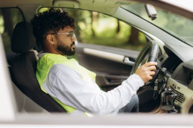 Şoför çelik yelek ve gözlük takıyor, direksiyon tutuyor, yolculuk sırasında yola odaklanıyor.