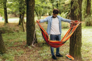 Bir adam yemyeşil bir parktaki bir ağaca renkli bir hamak bağlıyor. Rahatlamaya ve doğanın tadını çıkarmaya hazırlanıyor.