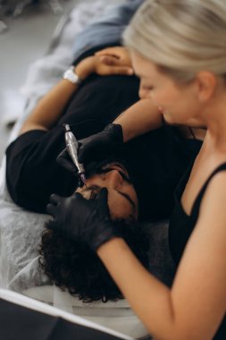 Güzellik uzmanı bir adamın kaşlarına kalıcı makyaj uyguluyor, profesyonel bir salon ortamında estetik dövme hizmeti veriyor.
