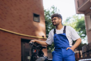 Mavi tulumlu neşeli teslimatçı, elektrikli scooterına yaslanıyor, gülümsüyor ve uzaklara bakıyor, yerleşim alanındaki teslimatlar arasında.