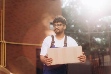 Güneşli bir yaz gününde kalabalık bir mahallede karton kutu taşıyan genç teslimatçı.