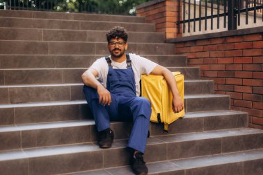 Kurye yemek siparişi verdikten sonra merdivenlerde dinleniyor. Yanında sarı yalıtılmış bir termal çantayla mola veriyor.