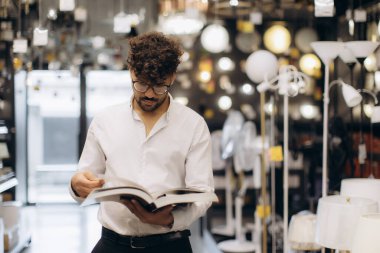 Beyaz gömlekli bir adam, iyi aydınlatılmış bir odada çeşitli aydınlatma armatürlerini gösteren bir kitap okur, bir odak atmosferi iletir ve iç dekorasyon için alışveriş yapar..