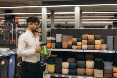 Bir mağaza koridorunda yeşil sulama kabını keşfeden ve taşıyan bir adam, istiflenmiş renkli saksıları incelerken, ev dekorasyonunu veya iç dekorasyon alışverişini örnek göstermektedir..