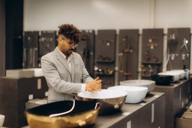 Bir adam modern bir hırdavat dükkanında çeşitli şık lavaboları inceliyor. Resim, çeşitli lavabo tasarımlarının estetik ve işlevselliğini gösteren ev donanımlarını seçme sürecini yakalıyor..