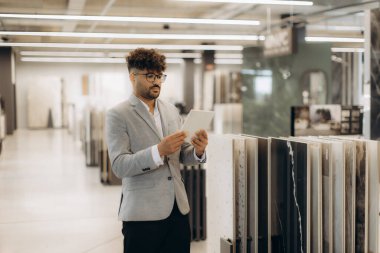 Bir iş adamı, modern bir galeride ürün görüntülerini incelerken dijital tablet bulundurur. Dekor, yenilik ve stili vurgulayan gösterişli, organize bir iç mekan içeriyor..