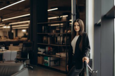 Modern bir ofiste profesyonel bir kadın, profesyonel ve çağdaş çalışma ortamlarını yansıtan, parlak mobilyalar ve endüstriyel tasarım unsurlarıyla çevrili, kendinden emin bir şekilde poz veriyor..