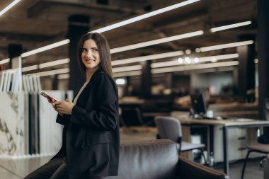 Rahat ve modern bir ofis ortamında akıllı telefon kullanırken gülümseyen kendine güvenen genç bir kadın. Ayarlar stil ve çağdaş dekora sahip profesyonel bir çalışma alanı gösteriyor.