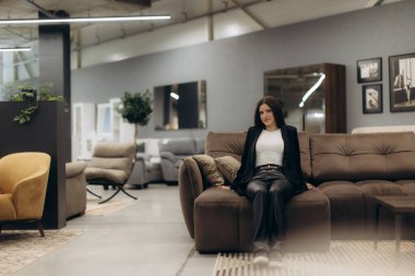 Modern bir koltukta modern bir mobilya mağazasında şık bir kadın oturuyor. Etrafında sandalyeler ve masalar gibi çeşitli mobilyalar var. Şık ve zarif bir ortam yaratıyorlar..