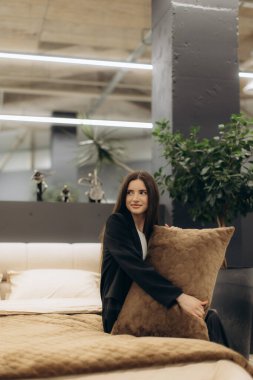 Genç bir kadın modern bir yatakta oturuyor, modern mobilya ve bitkilerle çevrili dekoratif bir yastığı kucaklıyor, iyi ışıklandırılmış ve şık bir odada rahat ve huzurlu bir atmosfer yaratıyor..