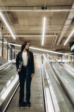 Profesyonel bir kadın, modern ofis ortamında kendine güvenen bir yürüyen merdivende duruyor, kararlılık ve entelektüelliği somutlaştırıyor. Ticari, kurumsal veya mesleki konular için ideal.