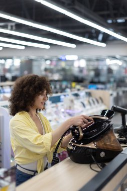 Sarı gömlekli bir kadın, çeşitli ürünlerin yer aldığı iyi ışıklandırılmış bir elektronik mağazasında modern bir demir alete bakıyor..