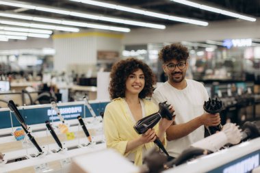 Neşeli bir çift, bir elektronik mağazasında saç şekillendirme cihazlarına, aksesuarlı fön makinelerine göz atar. Yeni tımar araçlarını keşfetmeye hevesli görünüyorlar..