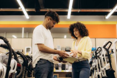 İki kişi modern bir elektronik mağazasında ev aletlerini incelerken görülüyor, özelliklerini tartışıyorlar. Mağazanın çevresi çeşitli ürünler ve modern ışıklandırmaların sıralarını vurguluyor.