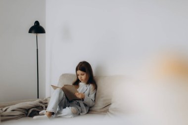 Kanepede oturup kitap okuyan bir kızın huzurlu bir sahnesi. Etrafı minimalist ve rahat bir iç mekan ile çevrili. Hayal gücü ve tefekkür için ideal rahat bir atmosfer yaratıyor..