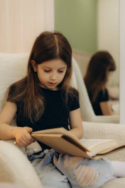 Genç bir kız, hafif bir koltukta rahatça oturur ve içeriğine dikkatle odaklanarak kitap okur. Ortam sıcak ve sakin. Merakını ve bilginin değerini gösteriyor..