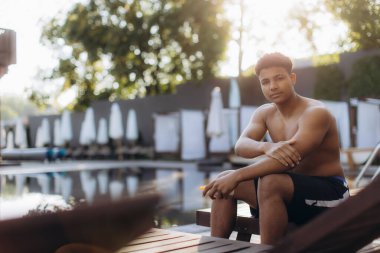 Genç adam havuz kenarında otururken koluna özenle güneş kremi sürüyor. Boş zamanlarında cilt bakımına ve güneş korumasına öncelik veriyor.