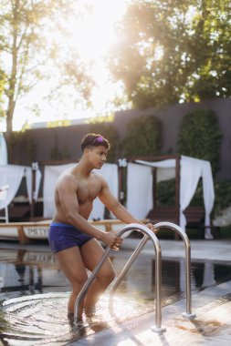 Eğitimden sonra metal merdiven kullanarak havuzdan çıkan kaslı genç sporcu gün batımının tadını çıkarıyor.