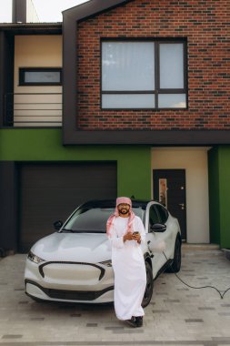 Modern bir elektrikli arabayı modern bir evin önünde şarj ederken ayakta duran ve akıllı telefon kullanan Arap bir adam, sürdürülebilirliği kucaklıyor.