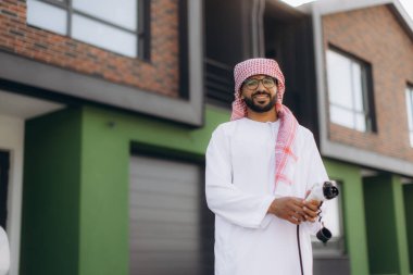 Geleneksel tobe ve ghutra giymiş gülümseyen Arap adam, dışarıda duruyor, modern bir evin önünde bir şarj aleti tutuyor.