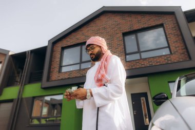 Çağdaş bir evin önünde duran ve geleneksel giysiler içinde para sayan bir Arap adam.