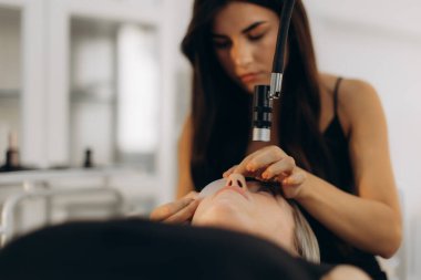 Güzellik salonunda güzellik ve göz makyajı üzerine çalışan bir güzellik uzmanı tarafından kirpik taktırılan bir kadın.