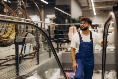 Bir mağazada yürüyen merdivenden inen iş kıyafetli bir adam, çeşitli ürünlerle perakende satış ortamı yansıtıyor.