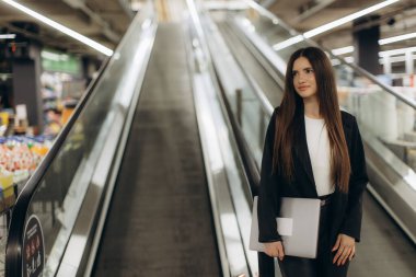 Profesyonel bir genç kadın yürüyen merdivende duruyor, elinde bir klasör, ticari bir çevrede tutku ve modern yaşam tarzını sembolize ediyor..