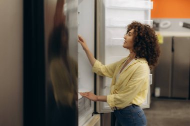 Sıradan sarı tişörtlü bir kadın bir elektronik mağazasında modern buzdolaplarını keşfediyor. Cihazı değerlendirirken memnun görünüyor, evden gerekli malzemeler için uygun bir alışveriş sahnesi öneriyor..