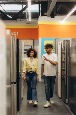 Bir çift buzdolabı seçeneklerine bakmak için bir elektronik mağazasına giriyor. Mağazada parlak ışıklar, modern cihazlar ve hoş bir alışveriş deneyimi sunan geniş bir yerleşim yeri bulunuyor..